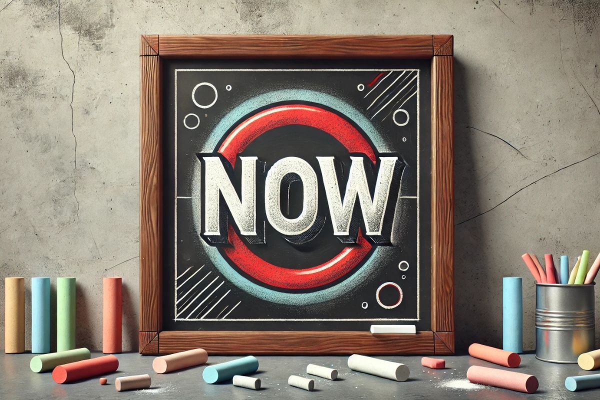 A wooden blackboard with the word "Now", surrounded by chalks.